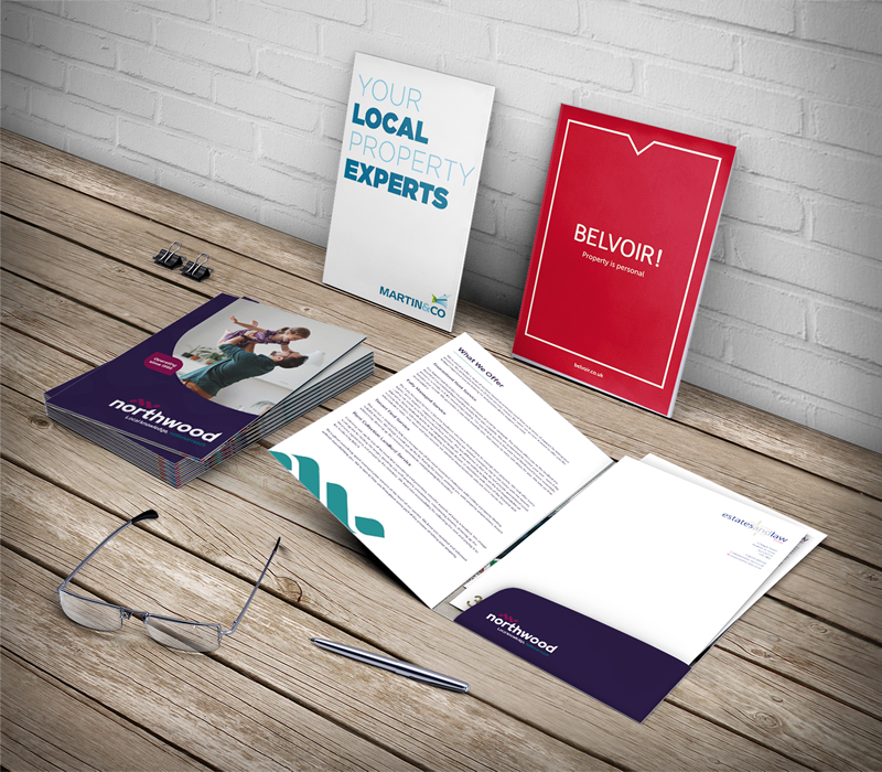 This image shows two brochures lead against a brick wall, one is red and the other is white. Infront of the white brochure are more brochures stacked up, these are purple. Besides this are documents in a file holder. Infront of these are glasses and a pen.
