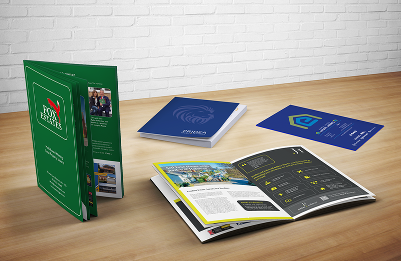 This image shows a green brochure opened up stood upright, next to this is another brochure with both pages showing. Behind this is a blue brochure lying on the desk besides this is a blue leaflet.